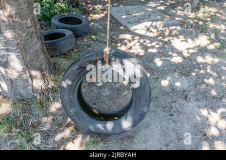 Oscillazione rustica fatta da una vecchia gomma dell'automobile appesa con le corde sull'albero contro il terreno asciutto, gomme nere e mele sul terreno, fondo sfocato, estate soleggiata Foto Stock