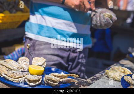 Ostriche fresche primo piano su piatto blu, servito tavolo con ostriche, limone e ghiaccio con un uomo che scopa un'ostrica. Cibo di mare sano. Foto Stock