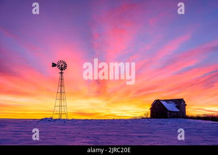 Una casa colonica in legno abbandonata e mulino a vento che si staglia con un brillante tramonto invernale nella rurale Alberta meridionale Foto Stock