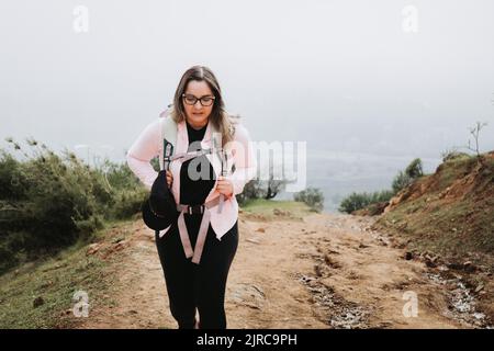 Stanco giovane latino più dimensione donna con zaino su, arrampicata su una collina e trekking in un bellissimo paesaggio. Foto Stock