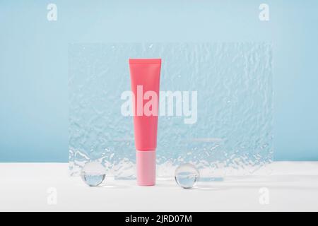 Tubo in plastica rosa crema cosmetica senza marchio mockup su sfondo blu con eleganti puntoni, palline di vetro e piastra acrilica. Lozione, celatore o bellezza Foto Stock