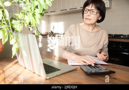 Una donna anziana bella e sofisticata che parla di videocomunicazione, lavora a casa con un notebook e fa calcoli su una calcolatrice. Foto Stock