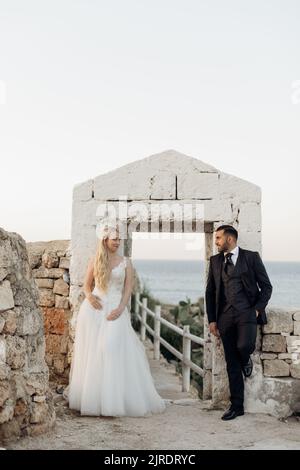 Ritratto di beatitudine coppia di nozze in piedi vicino ad arco antico. Giovane donna sposa guardando lo sposo in tuta. Verticale. Foto Stock