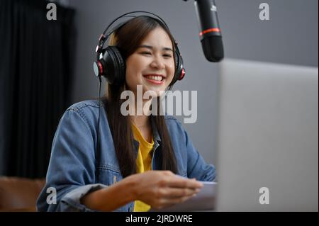 Allegro e felice giovane padrone di casa di radio online o freelance asiatico nel suo studio, ridendo e ama parlare con il suo ospite dalla chiamata Foto Stock