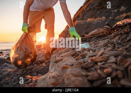 Giornata mondiale dell'ambiente. L'uomo volontario raccoglie una bottiglia di plastica sulla spiaggia. Primo piano. Spiaggia selvaggia sullo sfondo. Il concetto di pulizia della costa Foto Stock