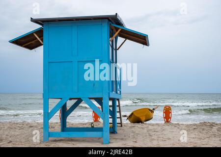 Un bagnino blu sulla spiaggia in una giornata nuvolosa. Una scialuppa di salvataggio gialla appoggiata al lato della casa. Foto Stock