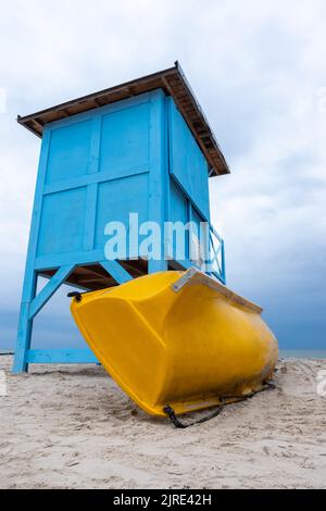 Un bagnino blu sulla spiaggia in una giornata nuvolosa. Una scialuppa di salvataggio gialla appoggiata al lato della casa. Foto Stock