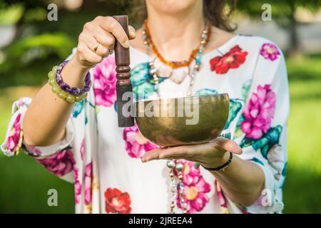 Dettaglio delle mani di una donna che tiene e suona una ciotola di canto tibetano durante una sessione di meditazione e terapia musicale in un parco. Foto Stock