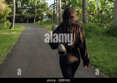 Vista posteriore di una giovane donna che porta un tappetino da yoga. Sulla strada per praticare lo yoga a Bali, sfondo verde Foto Stock