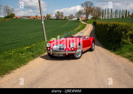 Una classica auto sportiva MGA si trova su una corsia di campagna in estate accanto a una lunga siepe con una casa colonica e alberi alti sullo sfondo Foto Stock