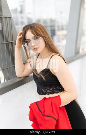 Elegante adorabile bella donna con i capelli lunghi indossare abito nero elegante e occhiali da sole in piedi e divertirsi in strada. Foto Stock