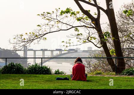 Giovane donna che si rilassa nei giardini Jardim do Palacio de Cristal a Porto, nel nord del Portogallo, con il fiume Douro e il ponte in lontananza. Foto Stock