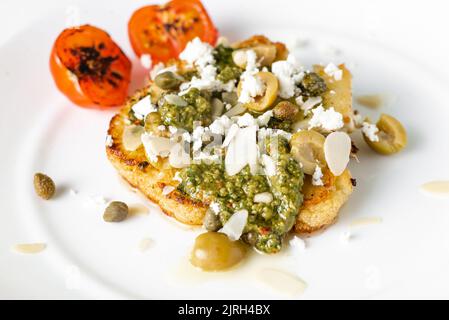 Bistecca di cavolfiore con spezie, salsa di chimichurri, fiocchi di mandorle, olive, pomodori ciliegini fritti e capperi su un piatto bianco. Cibo vegetariano. Foto Stock