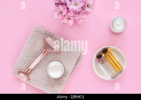 Crema in vaso, rullo facciale in cristallo di quarzo rosa, asciugamano e olio essenziale in bottiglie su sfondo rosa. Aromaterapia, auto-cura e cosmetici naturali Foto Stock