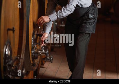 Enologo caucasico senior graybeard degusta vino rosso per verificarne la qualità e il sapore, accanto a un grosso barile di quercia Foto Stock