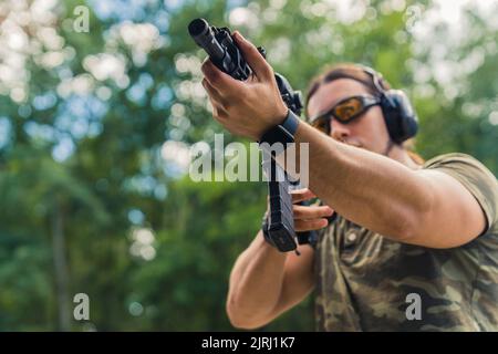 Uomo di mezza età caucasico dalla muscolosa fiducia in una t-shirt con motivi moro con fucile nero e loupe. Sfondo verde sfocato. Giorno al poligono di tiro. Foto di alta qualità Foto Stock