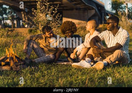 Gruppo multietnico di buoni amici che si godono il loro picnic estivo in natura mentre si siedono sull'erba vicino al fuoco, bevendo birra, parlando e guardando il tramonto. Foto di alta qualità Foto Stock