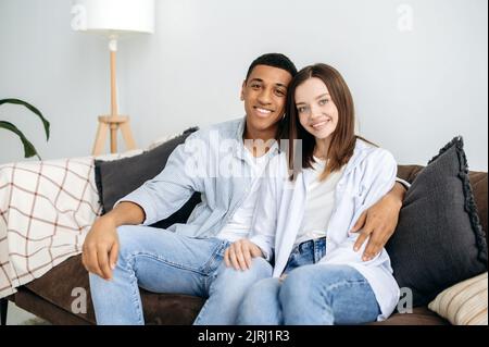 Coppia positiva felice sposata multirazziale innamorata, donna caucasica e uomo ispanico, seduto sul divano a casa in soggiorno, abbracciando, guardando la macchina fotografica, sorridendo, passare il tempo insieme Foto Stock