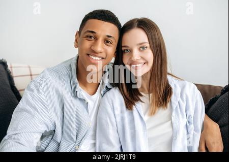Ritratto di una giovane coppia sposata felice. Coppia multirazziale positiva innamorata, donna caucasica e uomo ispanico, seduto sul divano a casa in soggiorno, abbracciando, guardando la macchina fotografica, sorridendo Foto Stock