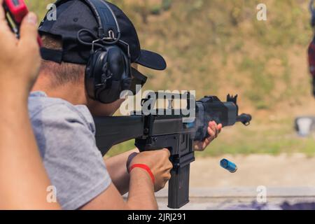 08.07.2022 Varsavia, Polonia uomo caucasico irriconoscibile in t-shirt grigia, berretto da baseball nero e cuffie protettive puntate con fucile nero. Gara di tiro. . Foto di alta qualità Foto Stock