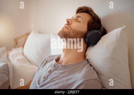 Rilassati, medita e riposa l'uomo ascoltando la musica con le sue cuffie wireless mentre ti rilassi sul letto a casa. Rilassato e tranquillo maschio sognare Foto Stock