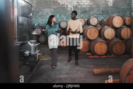 Cantina, magazzino e lavoratore di cantina insegnare un uomo sul vino mentre sta scrivendo note. Enologo che cammina in una distilleria con botti di legno Foto Stock