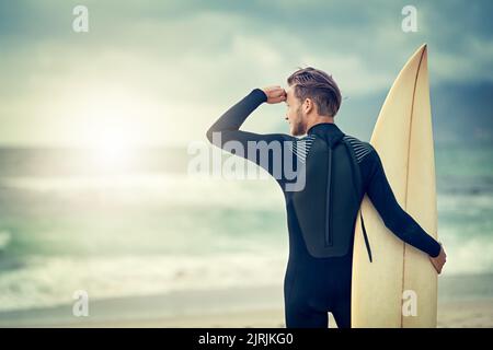 Scannerizzare il mare per una pausa perfetta. Un giovane surfista rilassato che guarda le onde mentre tiene la tavola da surf in spiaggia. Foto Stock