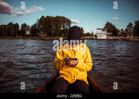 Giovane donna in impermeabile giallo e cappello seduta su una barca e guardando il telefono Foto Stock