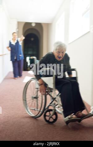 Prendimi se puoi. Una donna anziana maliziosa che usa la sua sedia a rotelle per scappare dalla sua infermiera in una casa di vecchiaia. Foto Stock
