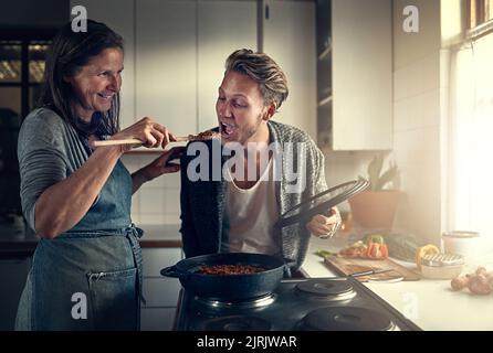 Cibo solo una mamma può cucinare. Madre che dà al figlio adulto un assaggio di quello che sta cucinando in cucina. Foto Stock