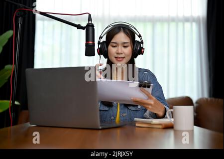 Affascinante donna asiatica che registra un podcast sul suo computer portatile con cuffie e microfono. Podcaster femminile che crea podcast audio dal suo studio di casa. Foto Stock