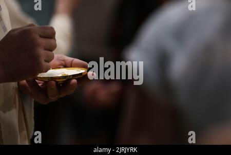 Il mistero della fede. Primo piano di un sacerdote cattolico che serve la messa e il pane della Santa eucaristia in comunione. Sfondo scuro ma host illuminato Foto Stock