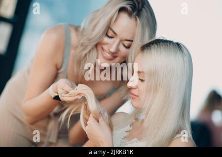 Parrucchiere femmina che fa estensioni di capelli a giovane donna con capelli biondi in salone di bellezza. Prolunga professionale per capelli Foto Stock