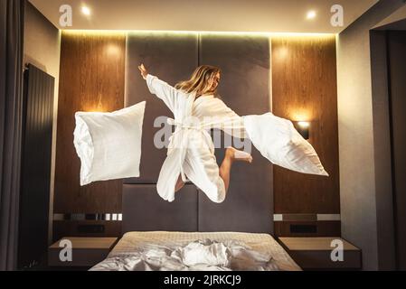 Donna che si diverte e che salta con l'accappatoio sul letto in camera d'albergo Foto Stock