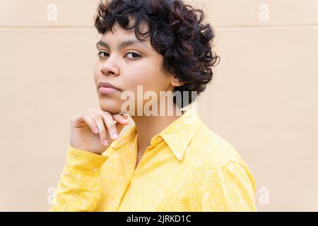 Ritratto di donna etnica raccolto con capelli corti ricci indossare camicia giallo brillante e mento toccante guardando delicatamente la macchina fotografica Foto Stock
