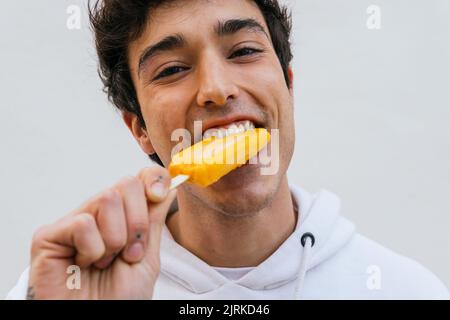 Felice giovane maschio mangiare dolce arancio ghiaccio lolly gustando il gusto su sfondo bianco Foto Stock