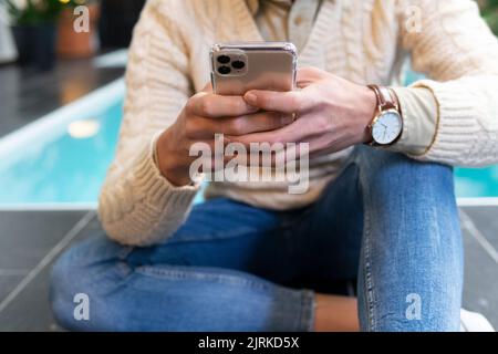 Anonimo giovane uomo ispanico in maglia maglione seduto a bordo piscina e navigare sul telefono cellulare Foto Stock
