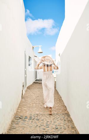 Vista posteriore irriconoscibile femmina a piedi nudi in una sundress bianca e cappello passeggiando su uno stretto sentiero tra case bianche sotto il cielo blu nelle Isole Canarie Foto Stock