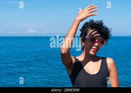 Allegra ragazza afroamericana con capelli ricci in piedi con il braccio in su mentre getta un'ombra sul suo volto contro l'oceano sotto il cielo blu Foto Stock