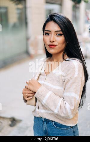 Giovane ragazza etnica con borsa che guarda la fotocamera sul passaggio piastrellato in città Foto Stock
