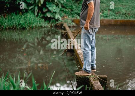 Vista laterale del pescatore irriconoscibile raccolto in abiti casual in piedi sul bordo di pietra nel fiume e la pesca di giorno Foto Stock
