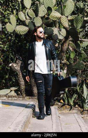 Chitarrista alla moda latino-americana maschile in giacca di pelle e occhiali da sole custodia con chitarra mentre si guarda lontano sulla scala contro le foglie di cactus Foto Stock