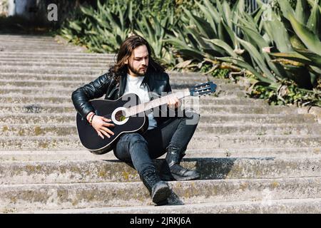 Musicista latino-americano alla moda seduto su scale urbane mentre suona la chitarra classica contro i cactus alla luce del sole Foto Stock