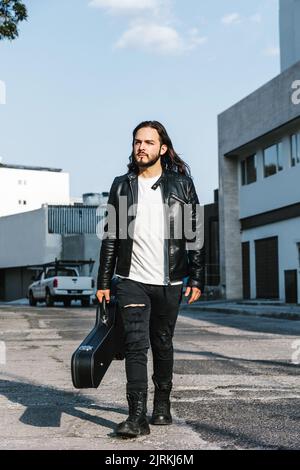 Chitarrista alla moda latino-americana maschile in custodia da trasporto giacca in pelle con chitarra mentre si guarda lontano sulla strada contro gli edifici Foto Stock