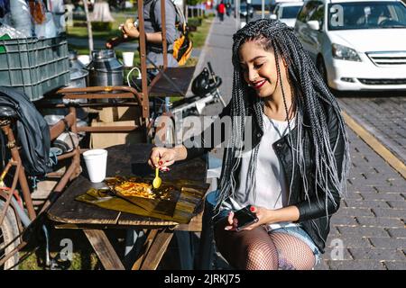 Femmina etnica tatuata con trecce afro e cucchiaio usa e getta seduto con gambe incrociate al tavolo della caffetteria con delizioso piatto in città Foto Stock