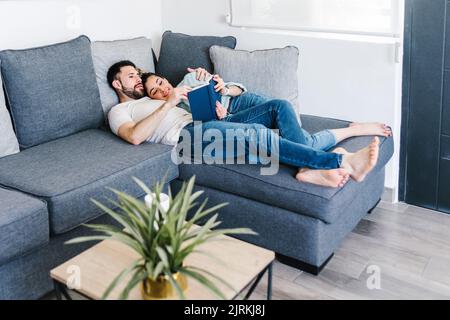 Tenera coppia latina che legge libro insieme mentre abbraccia e sdraiato sul divano nel soggiorno a casa Foto Stock