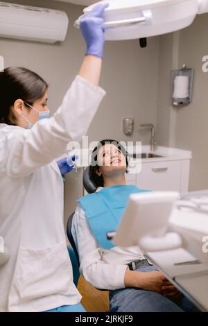 Vista laterale non riconoscibile stomatologo in uniforme vicino allegra paziente donna sdraiata sulla sedia in ospedale Foto Stock