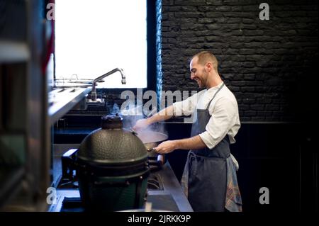 Giovane uomo allegro in grembiule grigio mescolando grande padella di metallo sulla cucina del ristorante Foto Stock