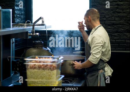 Giovane uomo in grembiule grigio assaggiando cibo da una grande padella di metallo sulla cucina del ristorante Foto Stock