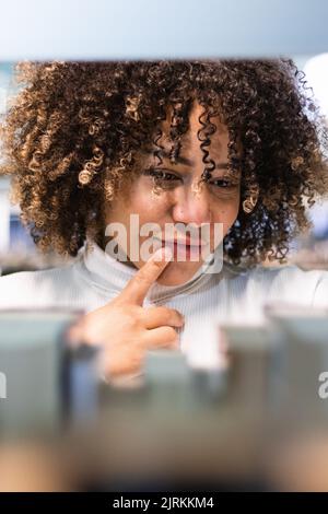 Studentessa afro-americana penosa con capelli ricci in piedi vicino alla libreria e raccolta libri mentre si passa il tempo in biblioteca universitaria Foto Stock